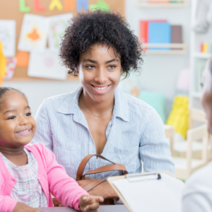 Black womanshort curly hair with a child next bedose her with a pink sweater talking to the teacher as part of their school routine.