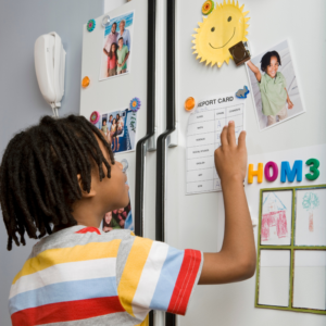 Neurodiverse Black child with medium length locs wearing a a blue, yellow, white, and red striped shirt in front of the refrigerator looking at the visual schedule.